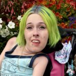Karli Drew. A white Disabled and visibly Queer person in a power wheelchair. She's notably pale with green eyes and unnaturally vibrant blue and green hair. They have a mermaid tank top on